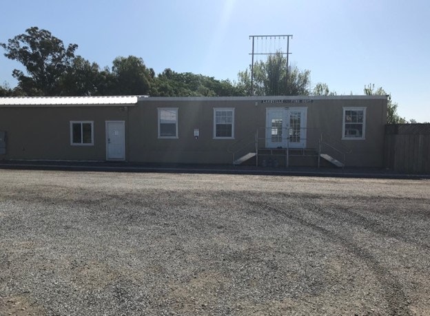 A single-story building with a central door, flanked by windows, under a clear sky, with a gravel lot in front.