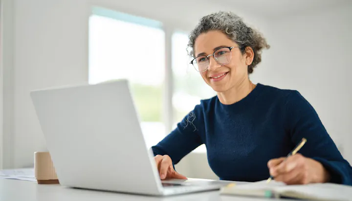 A woman with curly hair is smiling while working on a laptop and taking notes at a bright desk.