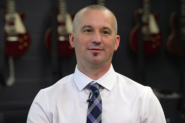 A man in a dress shirt and tie is in front of a backdrop featuring guitars, appearing engaged in conversation or thought.
