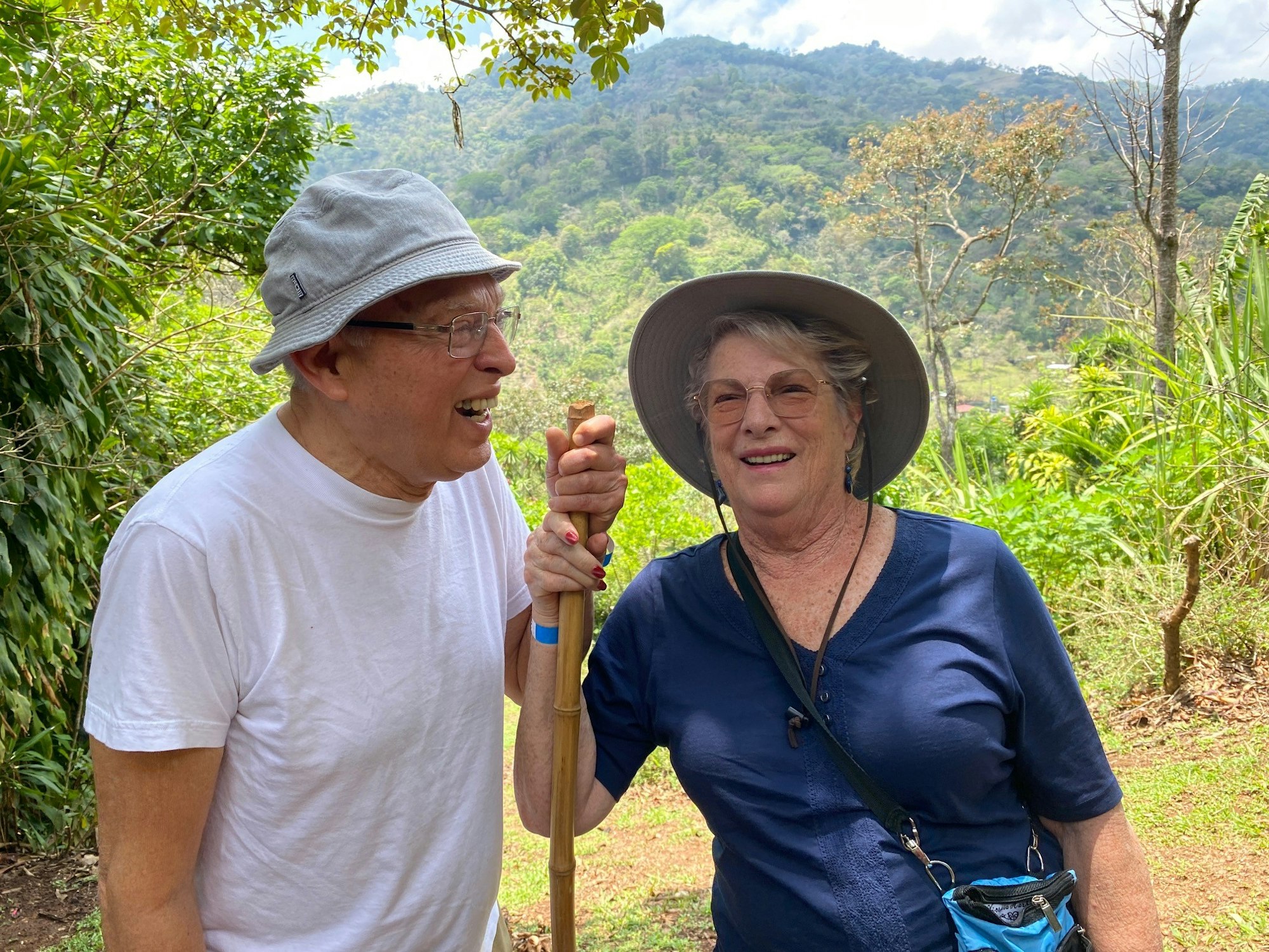 Two people smiling outdoors, wearing hats, with lush greenery and mountains in the background.