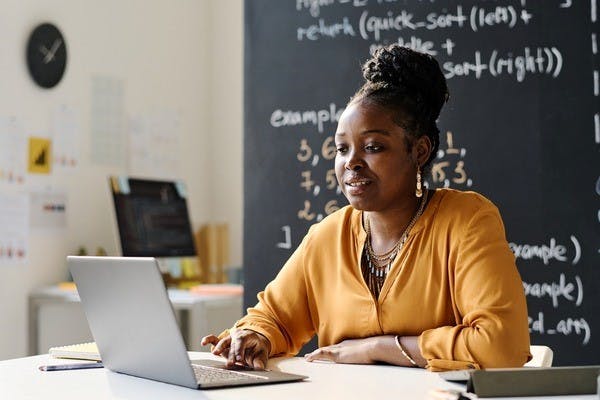 A person working on a laptop in an office with a blackboard in the background.