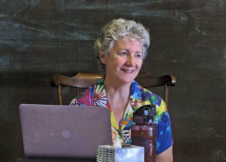 A person smiling, sitting in front of a laptop, wearing a colorful shirt, with a water bottle and tissue box on the table.