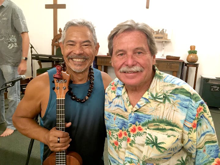 Two smiling men wear aloha shirts. One man has light gray hair and a mustache and holds a ukulele. Bill Dempsey has dark gray hair, a mustache and a bright shirt.