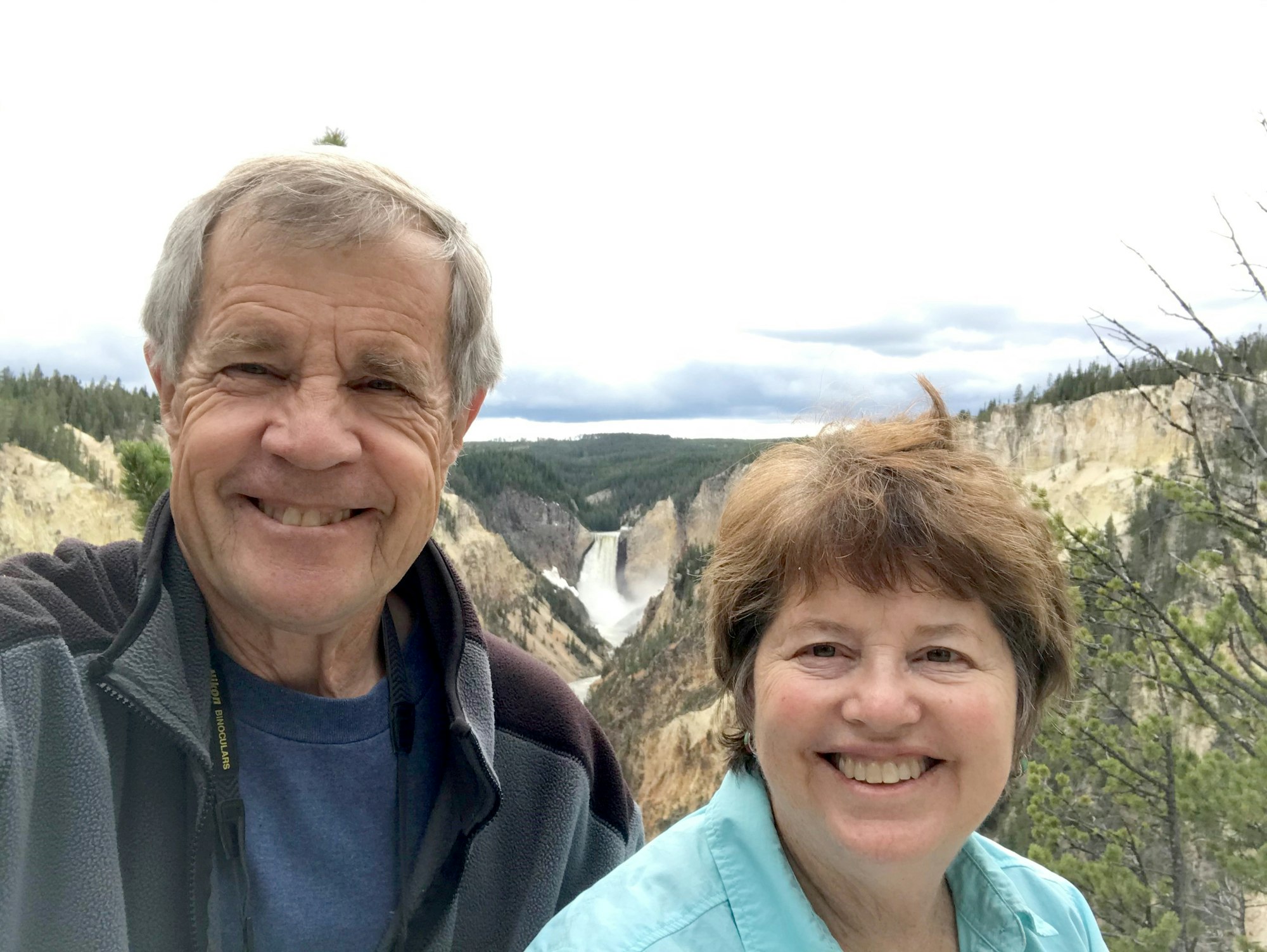 Rick and Janet Sitts are smiling, taking a selfie with a scenic waterfall and canyon in the background. Rick has gray hair and a gray jacket. Janet has brown hair and wears a light blue jacket.