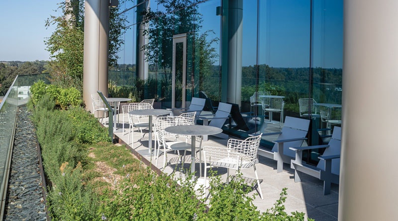 Modern patio with white chairs, tables, and greenery, beside glass walls overlooking a scenic view.