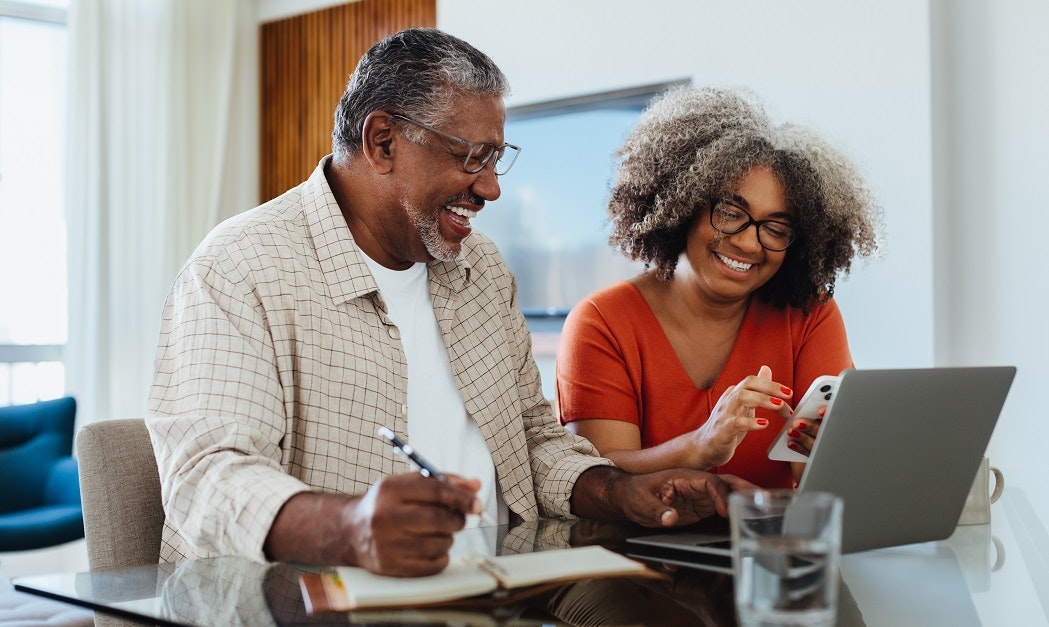 Two people smiling, looking at a laptop and a smartphone, engaging in a possibly productive or enjoyable activity together.