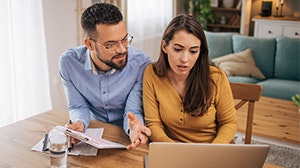 A man and woman are discussing something while working on a laptop at a table, showing expressions of focus and engagement.