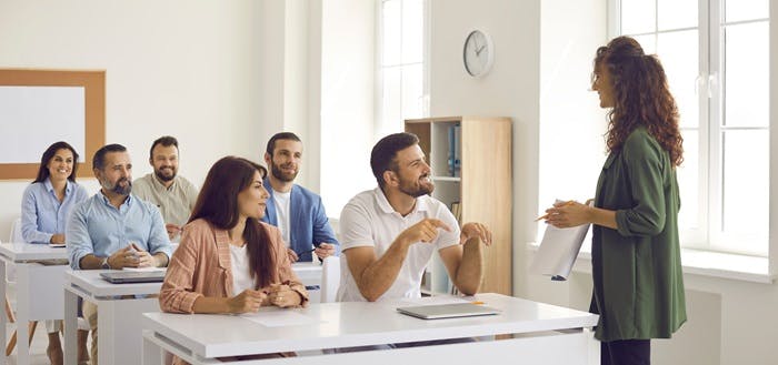 A group of six people sitting in a classroom, interacting with a teacher holding papers. The setting is bright and engaging.