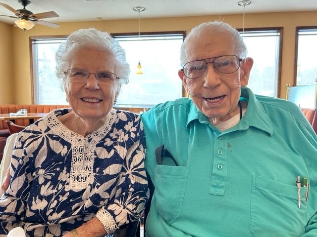 Mary Nelson has white hair and glasses and wears a blue and white floral shirt. Myron Nelson has white hair and glasses and wears a teal button up shirt.