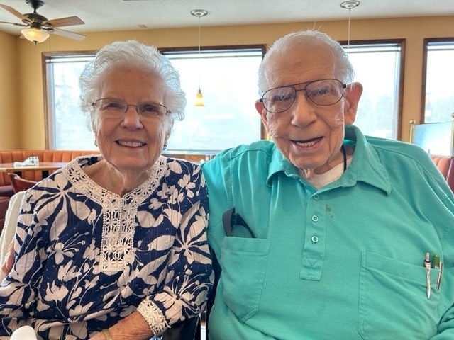 Mary Nelson has white hair and glasses  and wears a blue and white floral shirt. Myron  Nelson has white hair and glasses and wears a teal button up shirt.