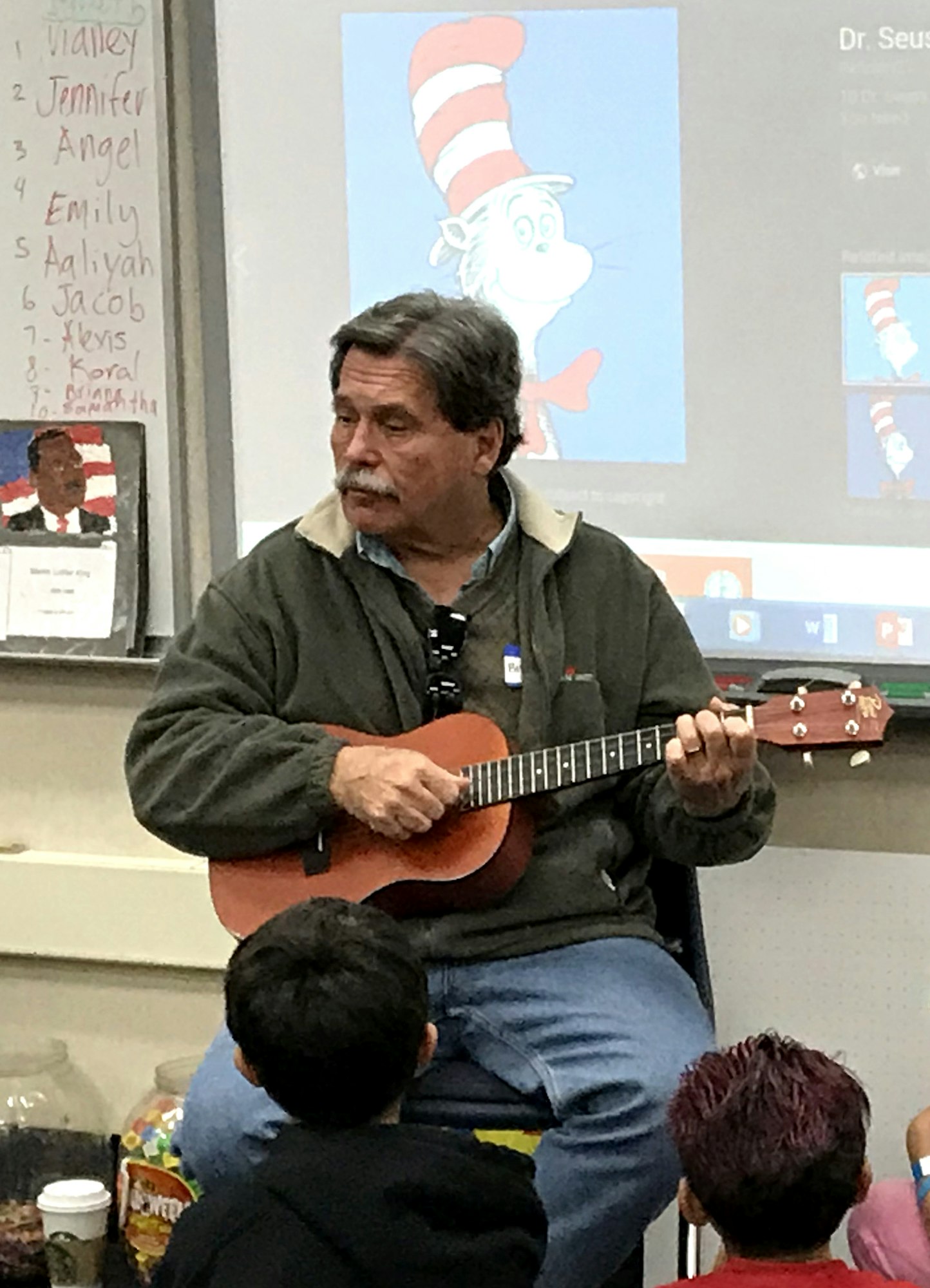 Bill Dempsey plays a ukulele for children in a classroom with a Dr. Seuss character on the screen.