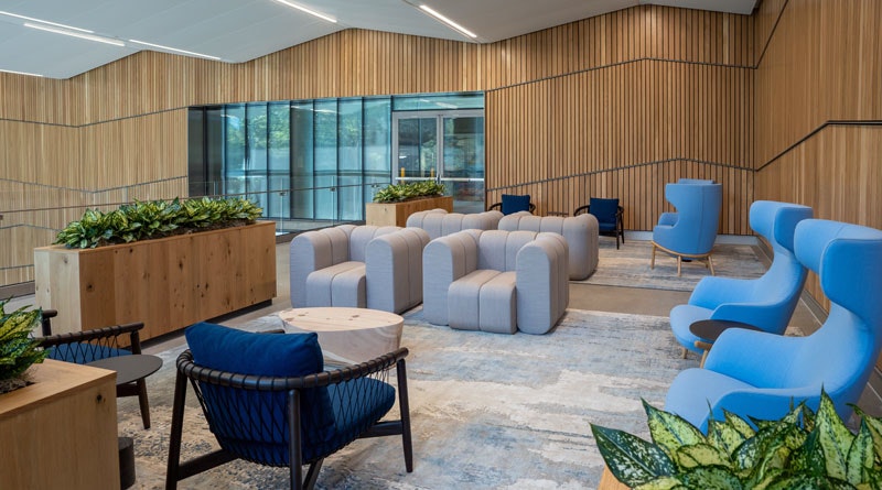 A modern lobby with gray and blue seating, wooden walls, plants, and natural light from large windows.