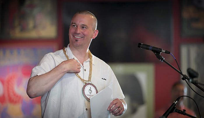 A smiling man in a white shirt stands at a microphone, wearing a large necklace and holding a paper, likely speaking at an event.