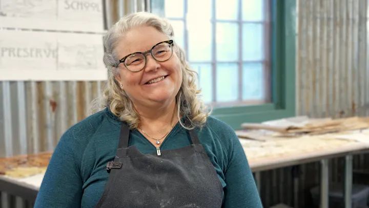 A smiling woman with curly hair, wearing glasses and an apron, stands in a workshop with a rustic background.