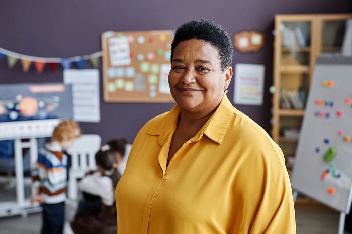 Woman in yellow shirt smiling, children in classroom background.