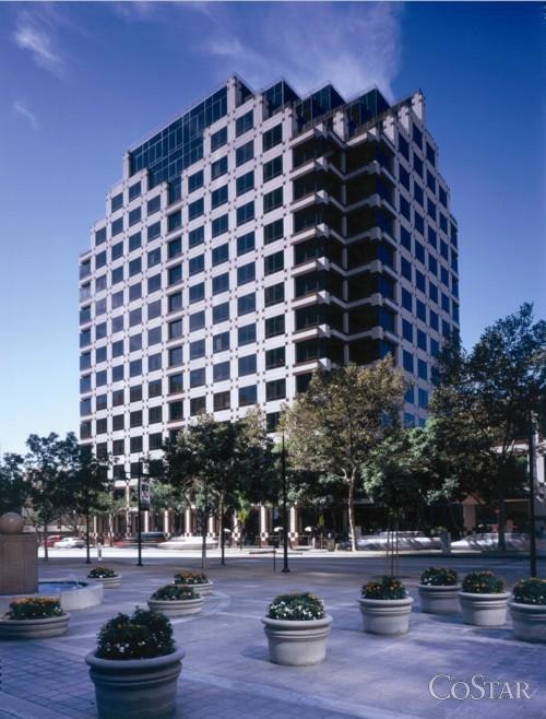 Modern high-rise building with reflective windows on a sunny day, viewed from a paved plaza with planters.