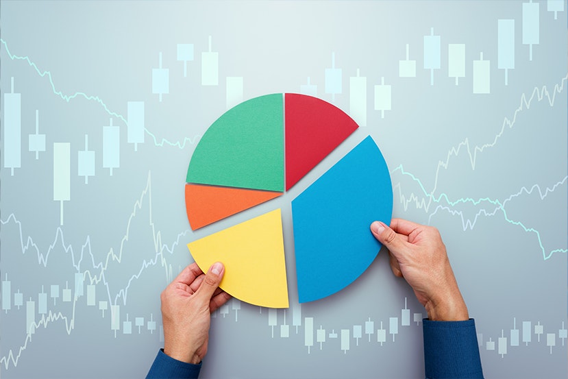 A pie chart with four colorful segments is held by hands against a background of financial graphs and candlestick charts.