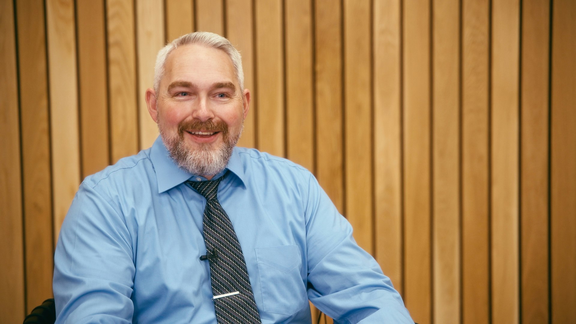 A bearded man in a blue shirt and tie sits in front of a wooden panel background, smiling.