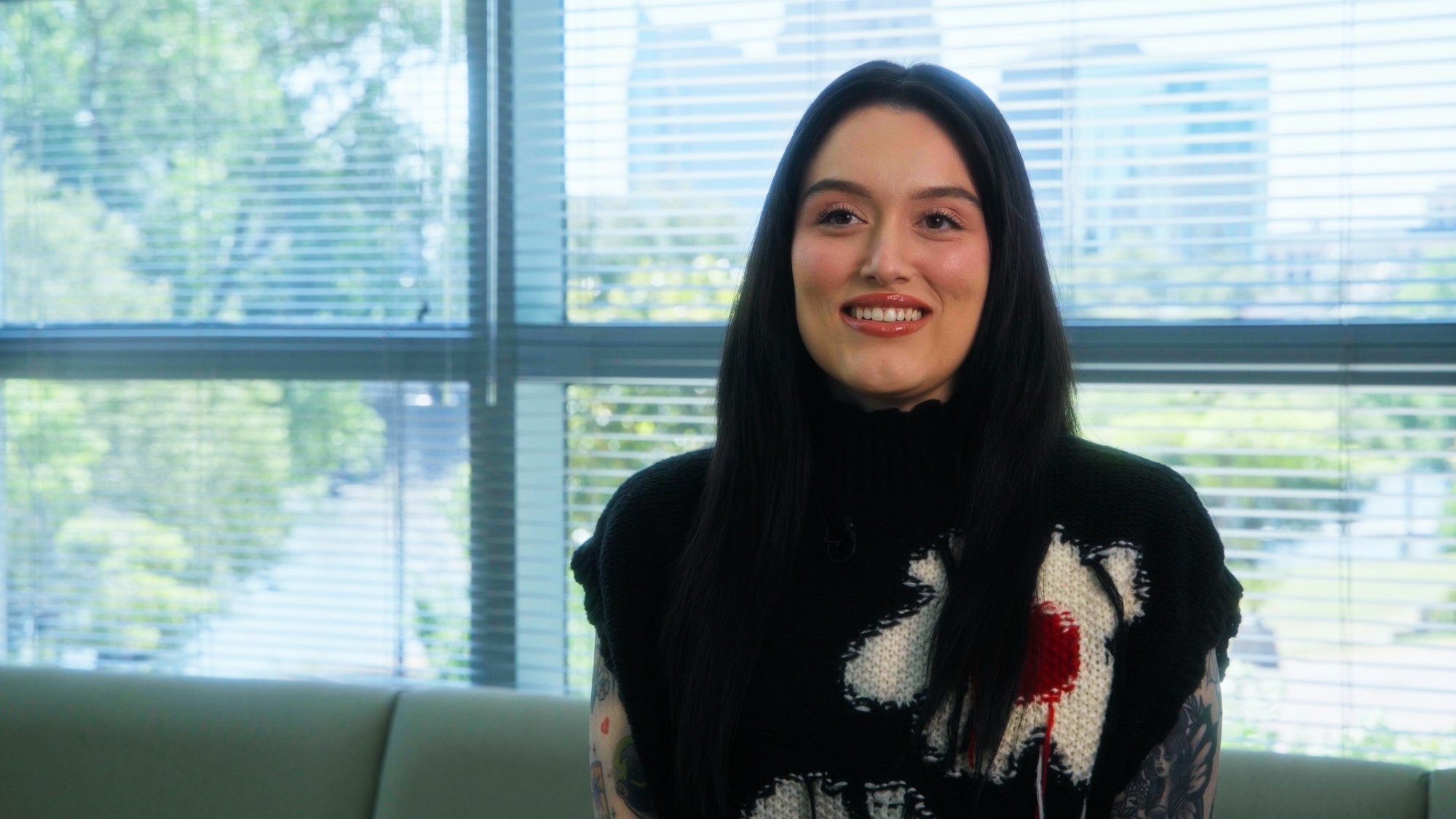 A person with long dark hair sits on a couch in front of a window with blinds, smiling.