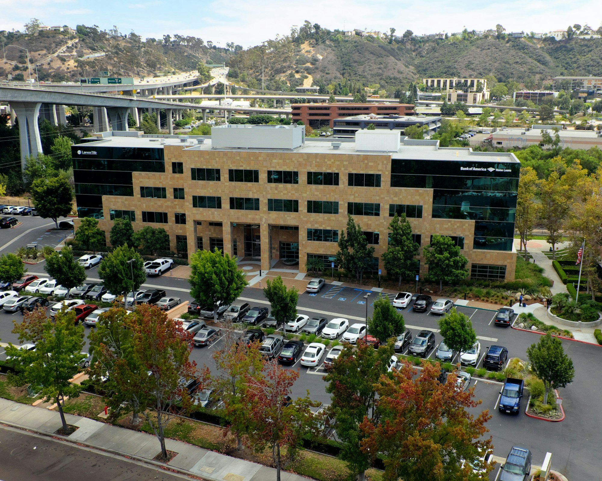 A multi-story building with a parking lot, surrounded by greenery, with a bridge and hills in the background.
