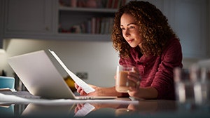 A woman with curly hair is sitting at a table, working on a laptop while holding a glass of drink and reviewing documents.