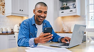 A smiling man is using a smartphone while sitting at a table with a laptop and documents in a modern kitchen.