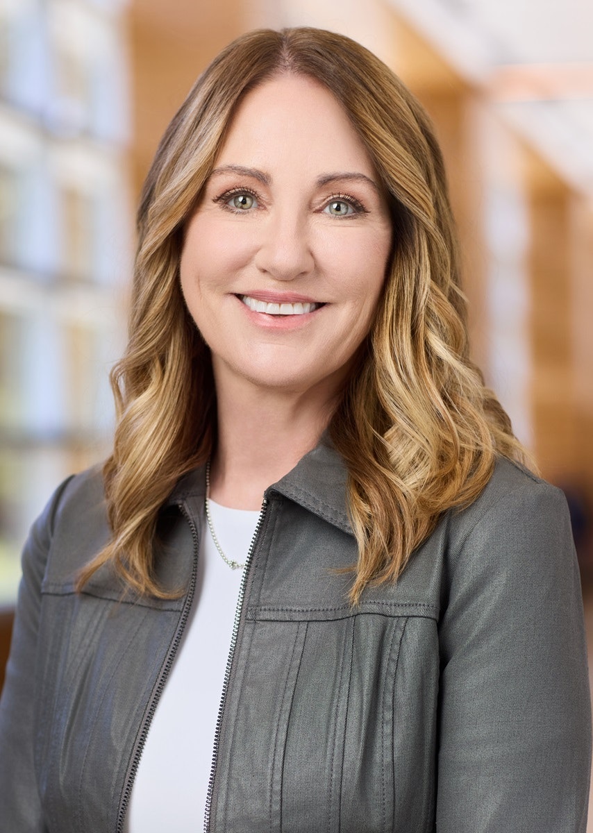 CalSTRS CEO Cassandra Lichnock, wearing a grey jacket and white shirt, smiling against a blurred indoor background.