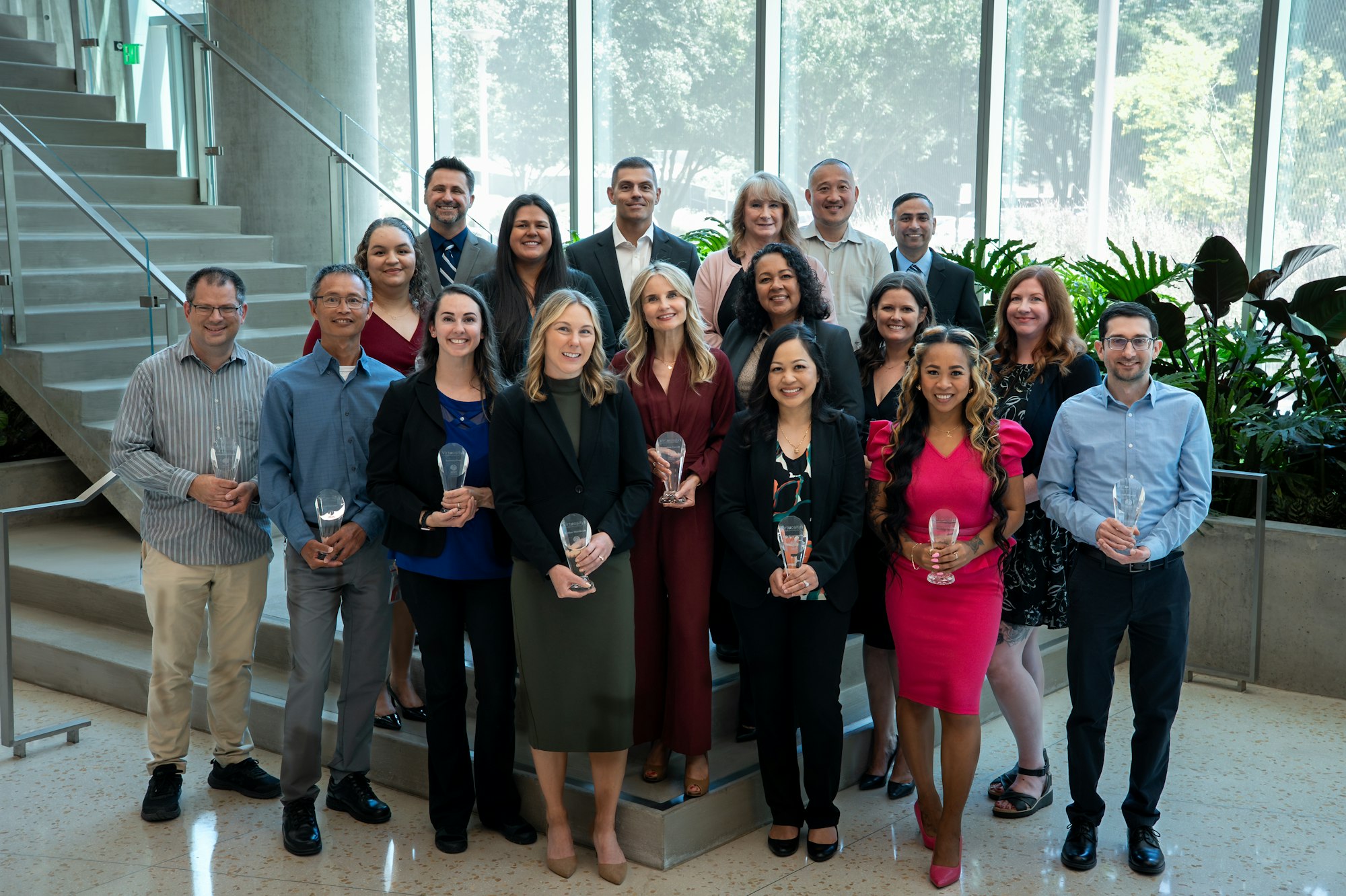 A group of people posing indoors, holding glass awards, with a staircase and large windows in the background.