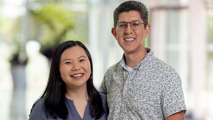 Two smiling people, possibly colleagues, standing together in a well-lit indoor setting with a blurred background.
