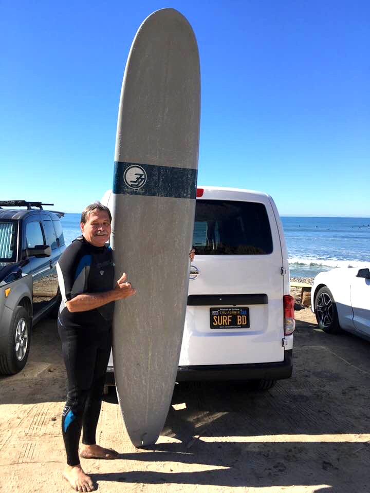 Bill Dempsey wears a wetsuit and stands with a surfboard by a beach parking lot.