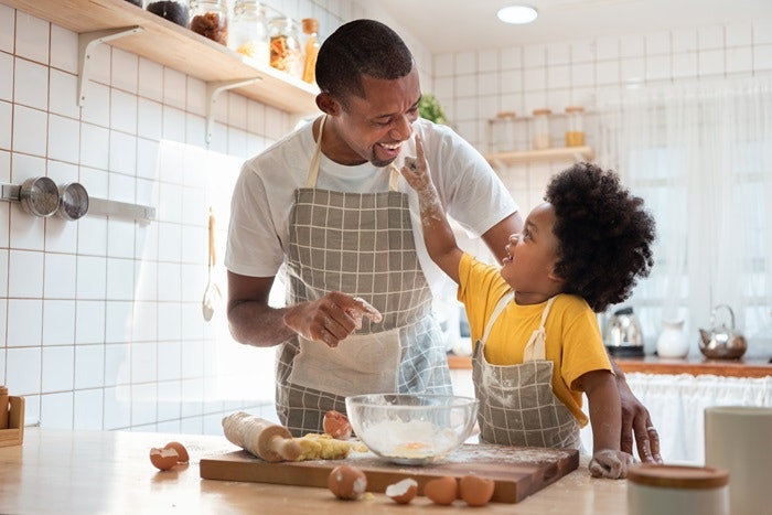 Father and child happily baking together in a kitchen, wearing aprons, with flour and ingredients on the counter.