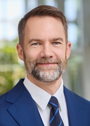 Jeff Zimmer in a suit and tie with a beard, smiling in an indoor setting with blurred greenery in the background.