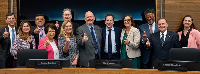 A group of smiling people giving thumbs up behind a conference table with nameplates.