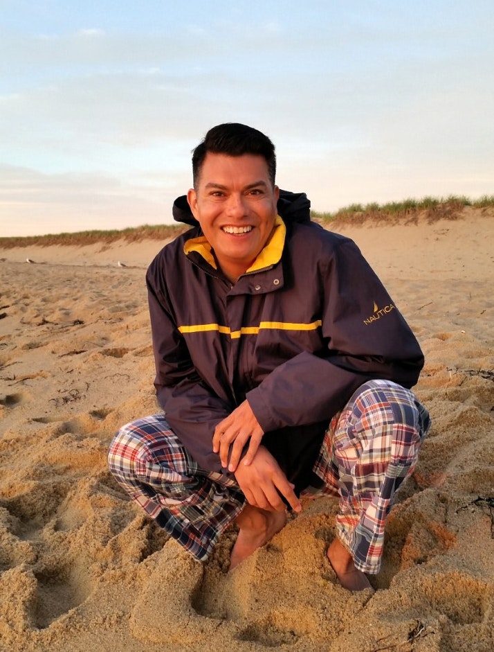 Francisco Laguna has short dark hair and is smiling. He crouches on sand at a beach with a cloudy sky behind him.