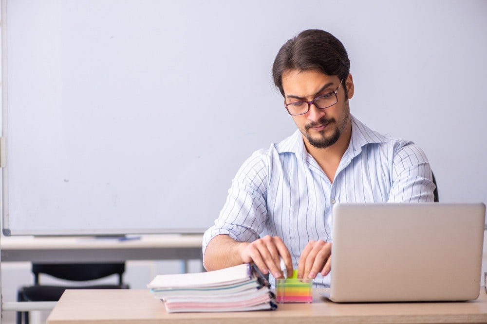 A man in a striped shirt sits at a desk, using a laptop, with colorful papers and a box of sticky notes in front of him.