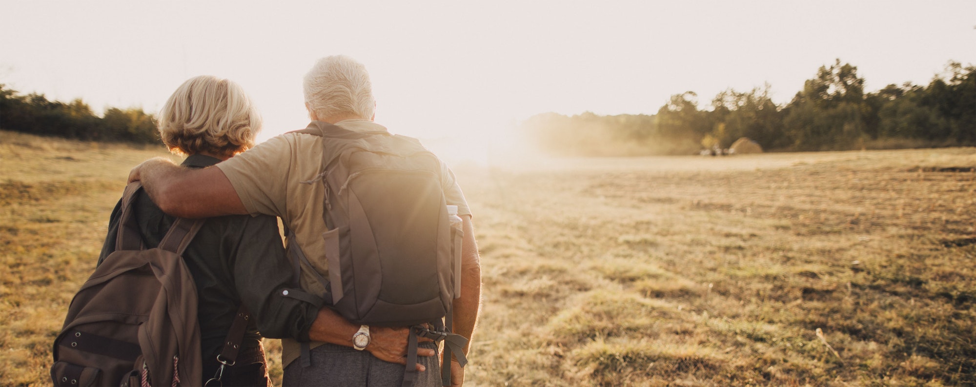 An older couple with backpacks embracing, looking at a field bathed in sunlight.