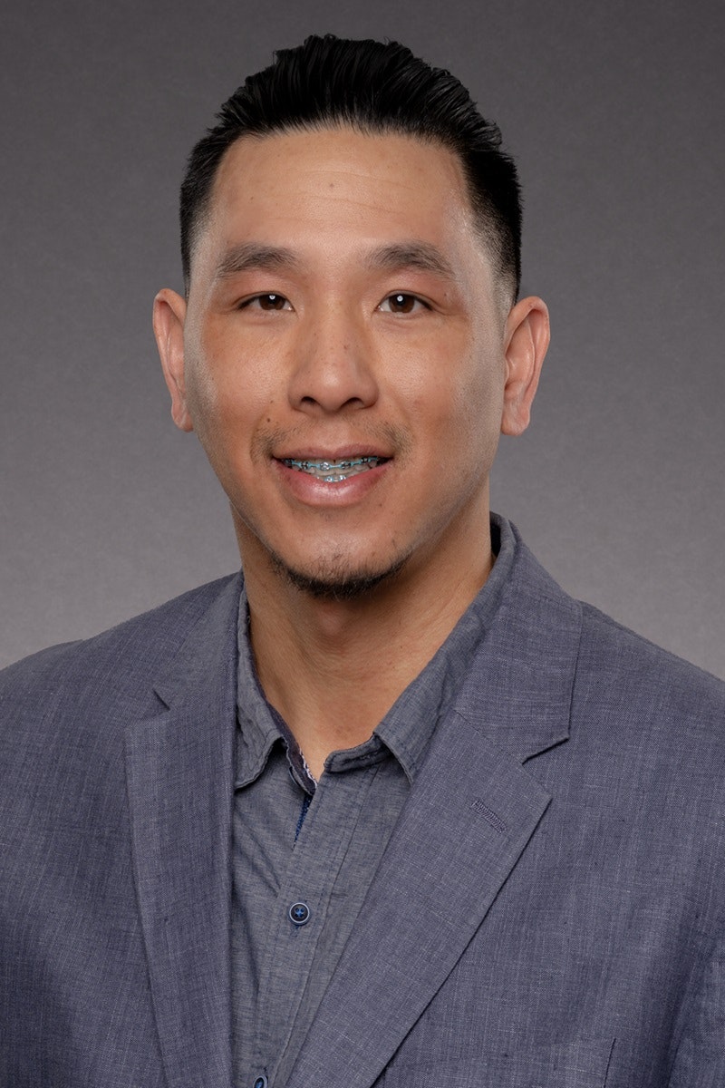 A person smiling, wearing a blue blazer and shirt, with a gray background.
