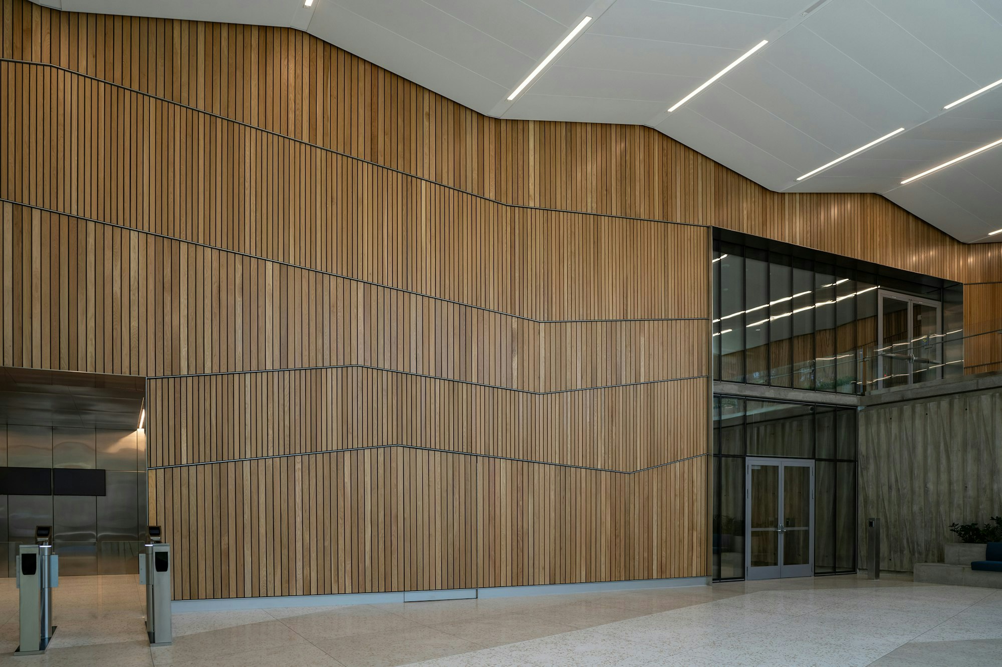 Modern lobby with wooden walls, geometric lighting, turnstiles, and glass facade.
