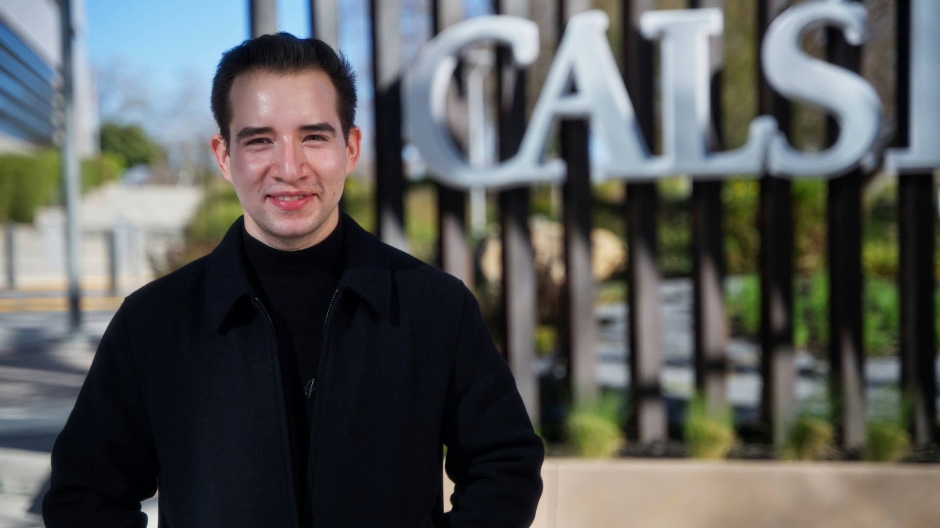 A person in a black coat smiles in front of a blurred background with large letters.