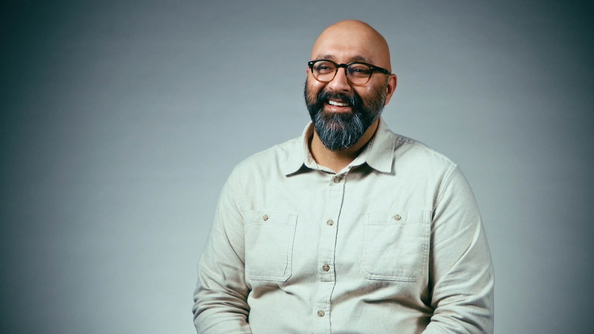 A man with glasses, beard, and a light-colored shirt is smiling against a plain background.