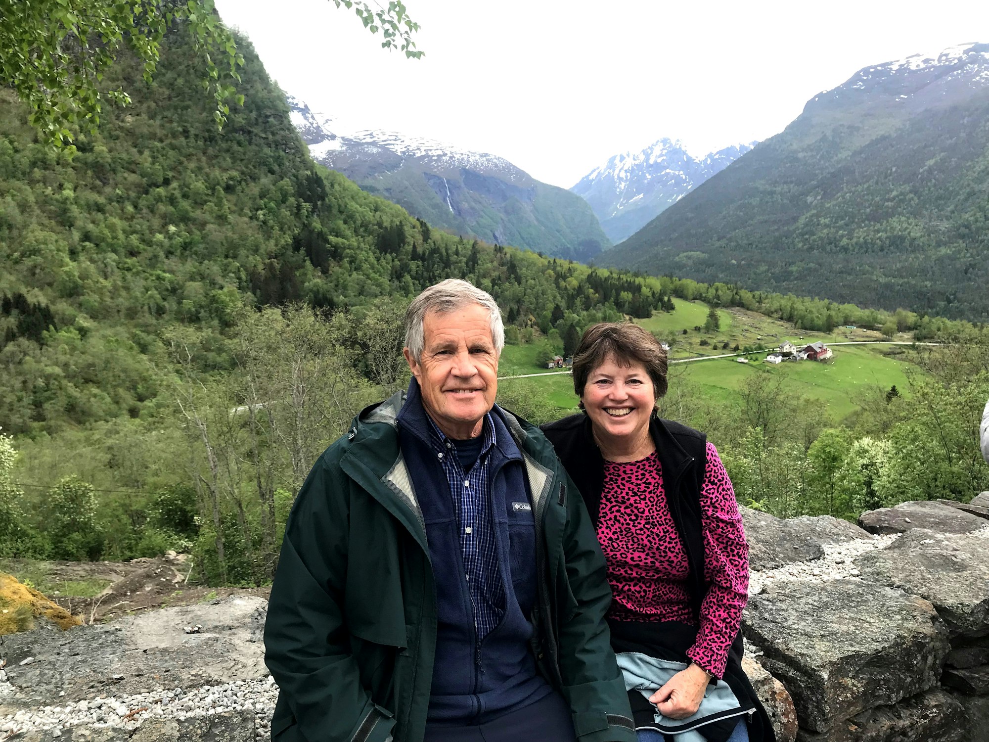 Rick and Janet Sitts pose before a scenic mountain valley with greenery and snow-capped peaks. Rick has gray hair and wears a dark jacket. Janet has brown hair and wears a magenta shirt.
