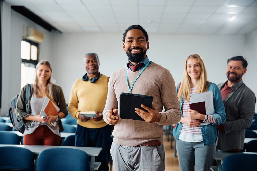 A diverse group of five people smiling in a classroom, holding books and a tablet.
