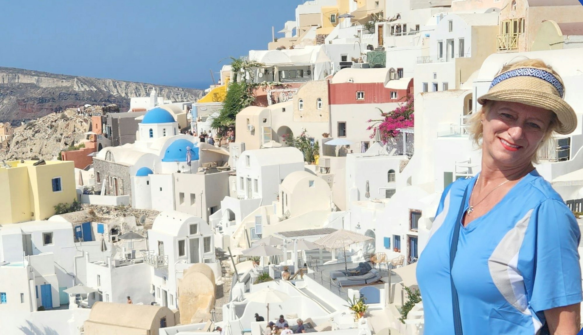 Kathy Cuevas has blond hair and wears a blue shirt and a straw hat. There is a city full of bright beige adobe houses and a body of water in the background.