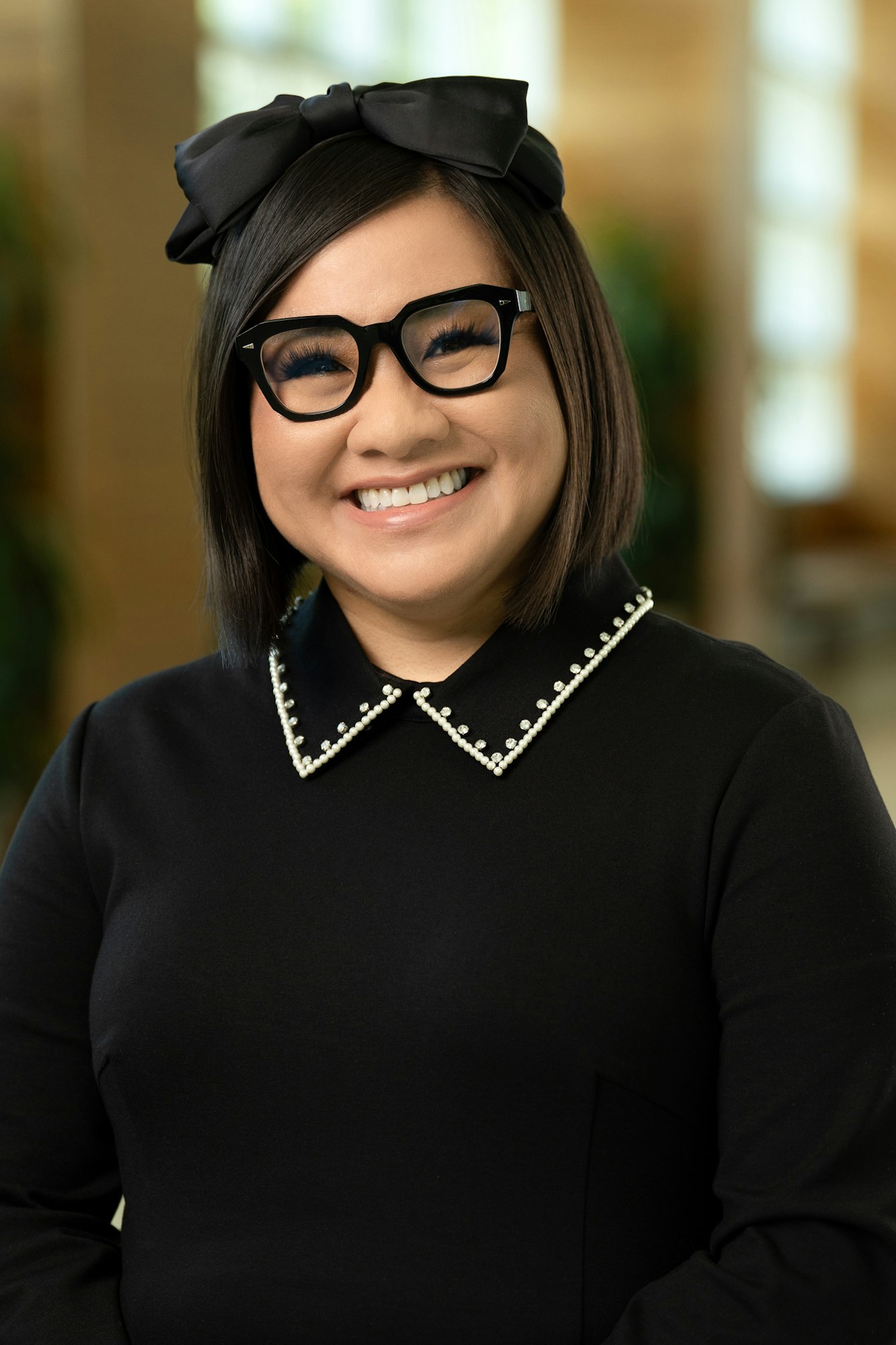 Christina Nguyen wearing glasses and a black outfit with a pearl-lined collar, with a black bow in their hair.