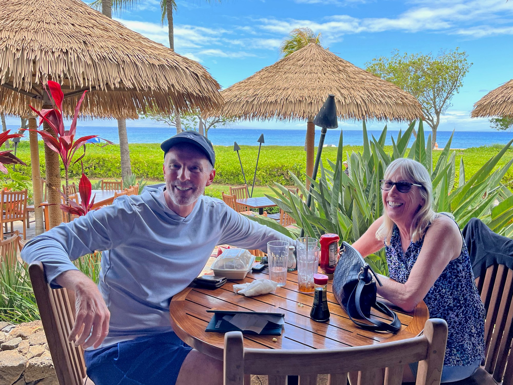 David Brown and his wife sitting at a wooden table outdoors with tropical plants and tiki umbrellas, enjoying a meal near the ocean.