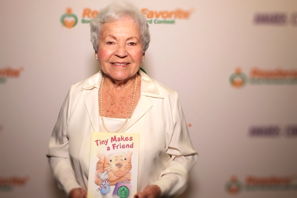 Eva Schmidler has short white curly hair and wears a white shirt. She is holding a children's book she wrote.