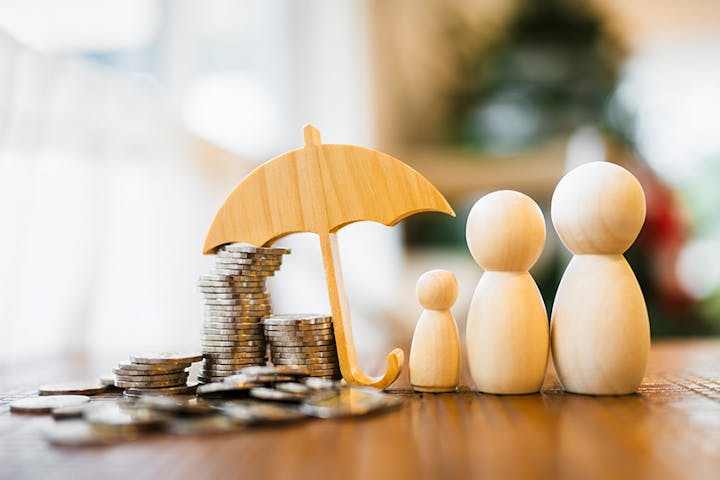 The image features wooden figures representing a family, an umbrella, and stacks of coins, symbolizing financial protection and savings.