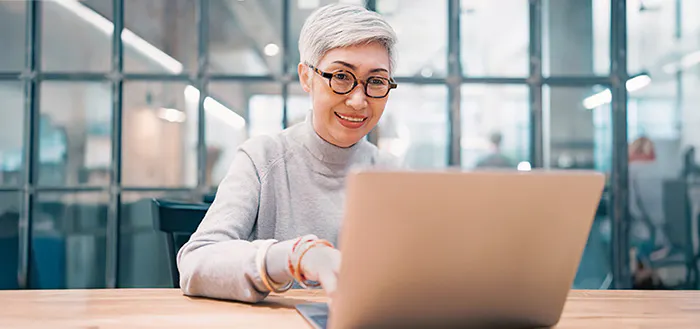 A smiling person with short gray hair and glasses works on a laptop in a modern office setting.