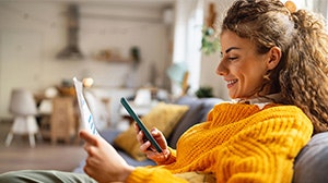 A person in a cozy sweater is sitting on a couch, smiling while using a tablet and a smartphone in a bright living space.