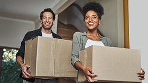 A man and a woman smile while carrying large cardboard boxes inside a house, suggesting a moving or unpacking activity.